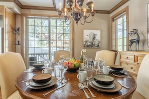 A dining room with a wooden table set for six, surrounded by chairs. A chandelier hangs from the ceiling, and there are windows with a view of trees. - Walk to Canyon Lodge | Noble Nest + Hot Tub/Sauna + 2-Car Garage (Mammoth)