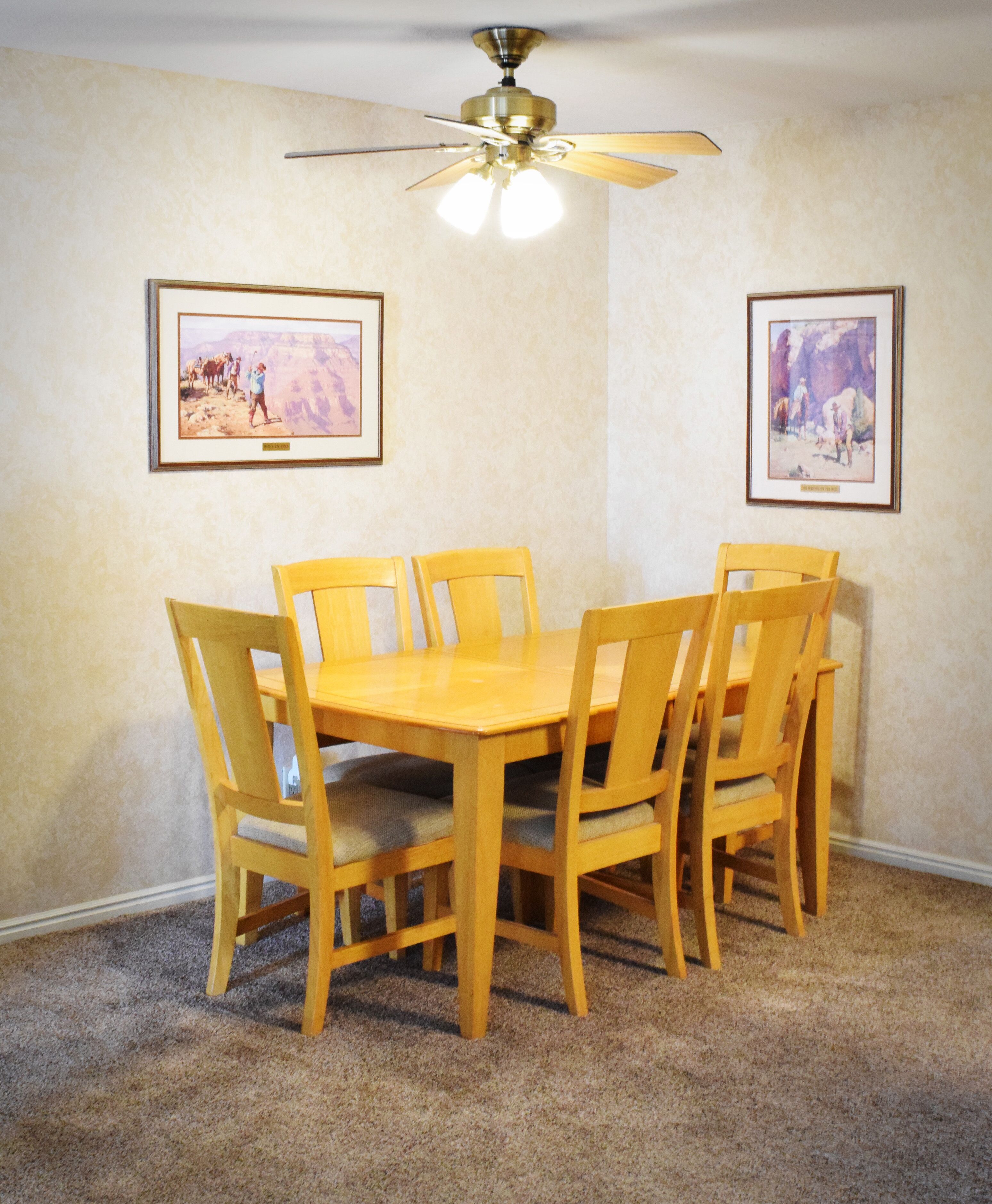 A dining room with a wooden table and chairs, a ceiling fan, and two framed pictures on the walls.