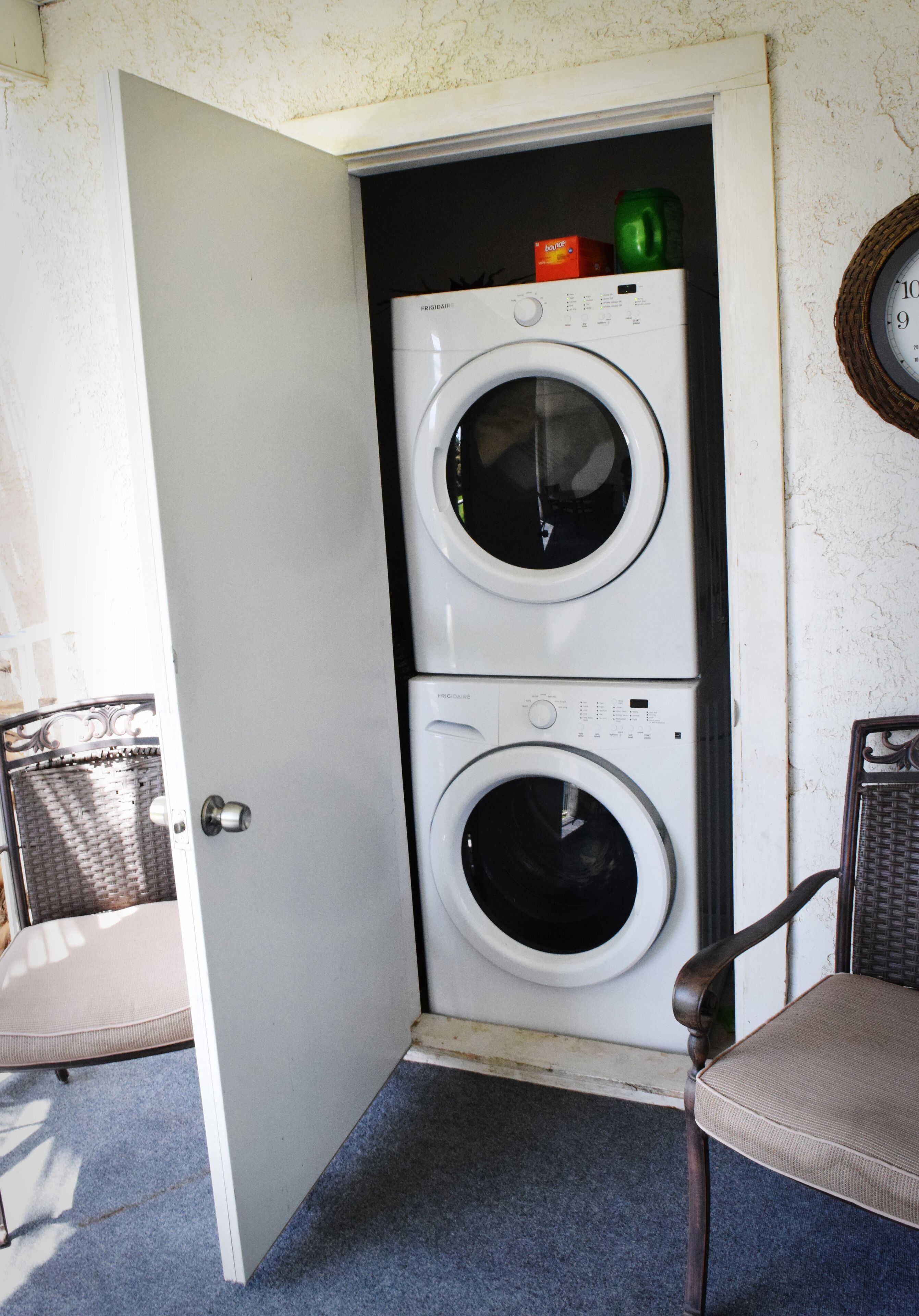 A laundry room with two stacked washing machines, a wicker chair, and a door leading to another room.