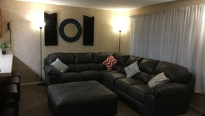 Ett vardagsrum med en hörnsoffa, en pall, ett soffbord, en lampa och en spegel som hänger på väggen.