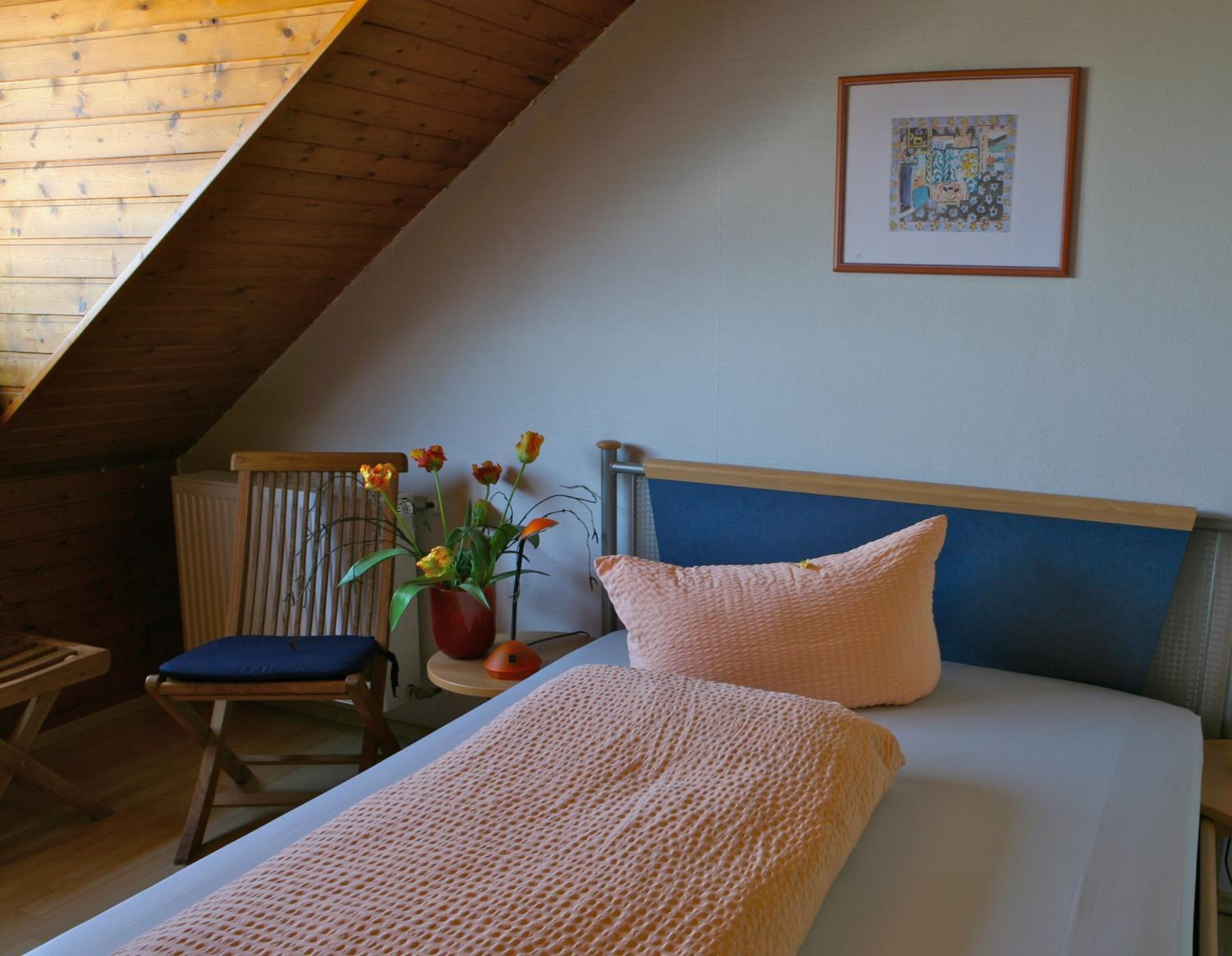 Un dormitorio con techo inclinado, una cama con cabecera azul, un cojín rosa, una silla azul y un jarrón con flores.