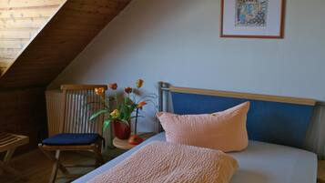 Un dormitorio con techo inclinado, una cama con cabecera azul, un cojín rosa, una silla azul y un jarrón con flores.
