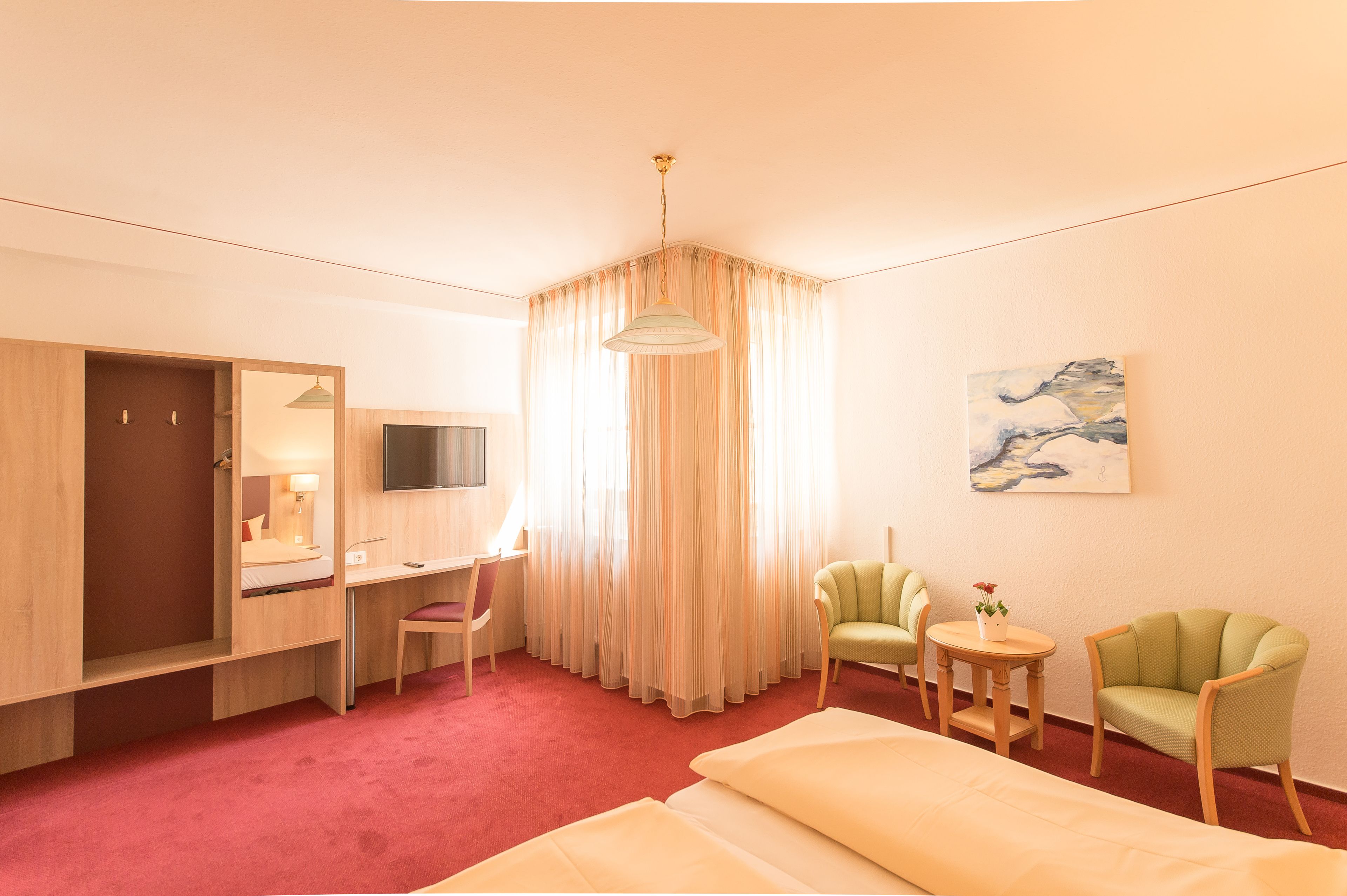 A hotel room with a bed, a desk, a chair, a small table, a mirror, a television, and a painting on the wall.