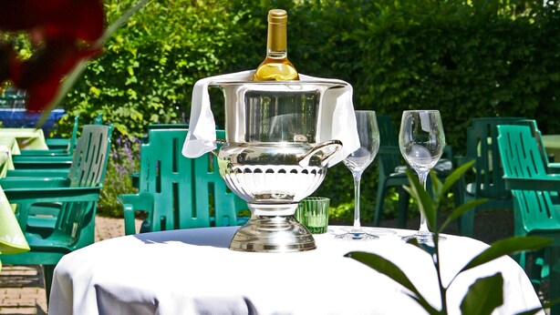Una mesa con un decantador de plata y vasos, colocada al aire libre con sillas verdes y un mantel blanco.