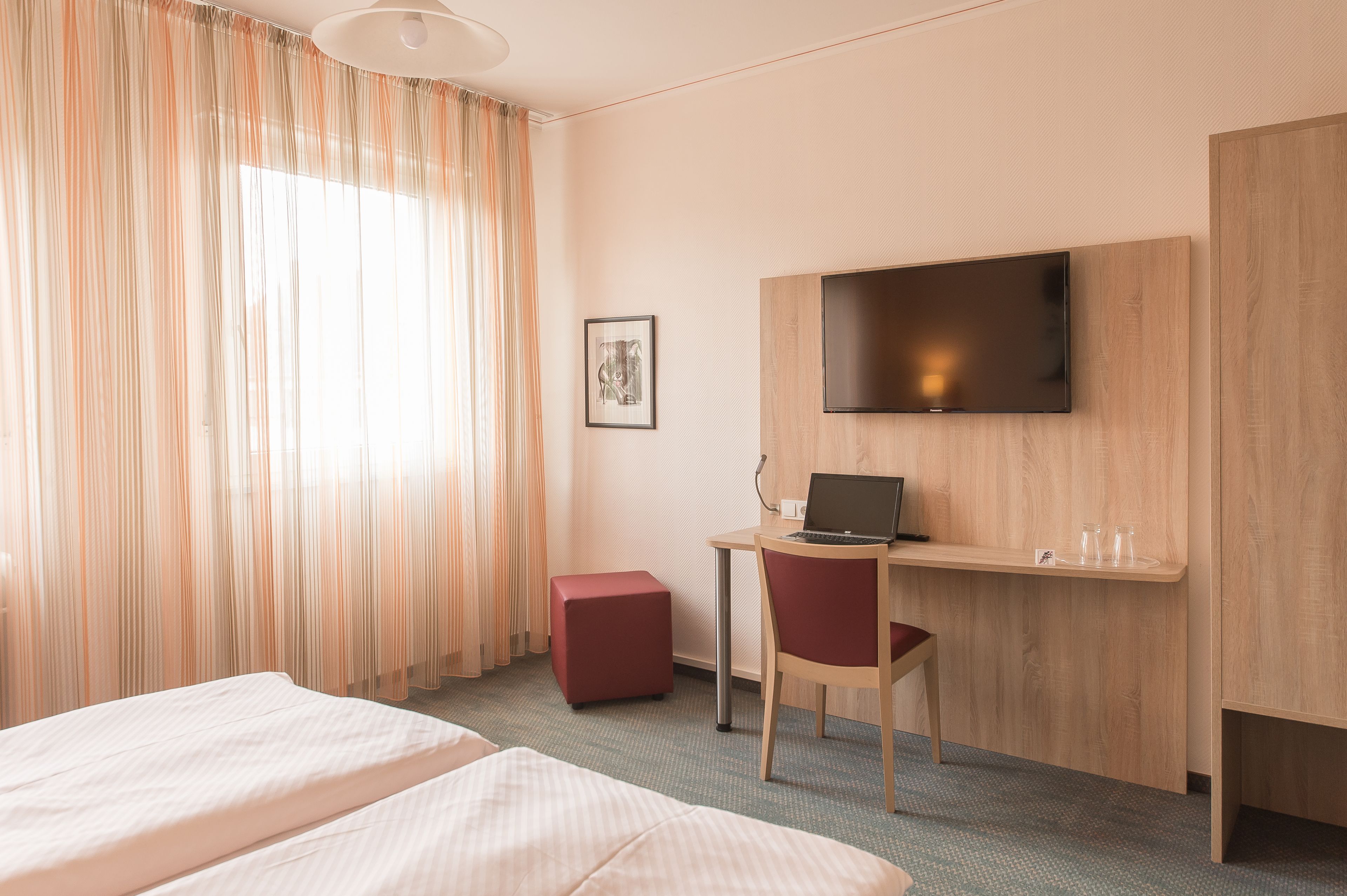 A hotel room with a bed, a desk with a laptop, a TV, and a red ottoman.
