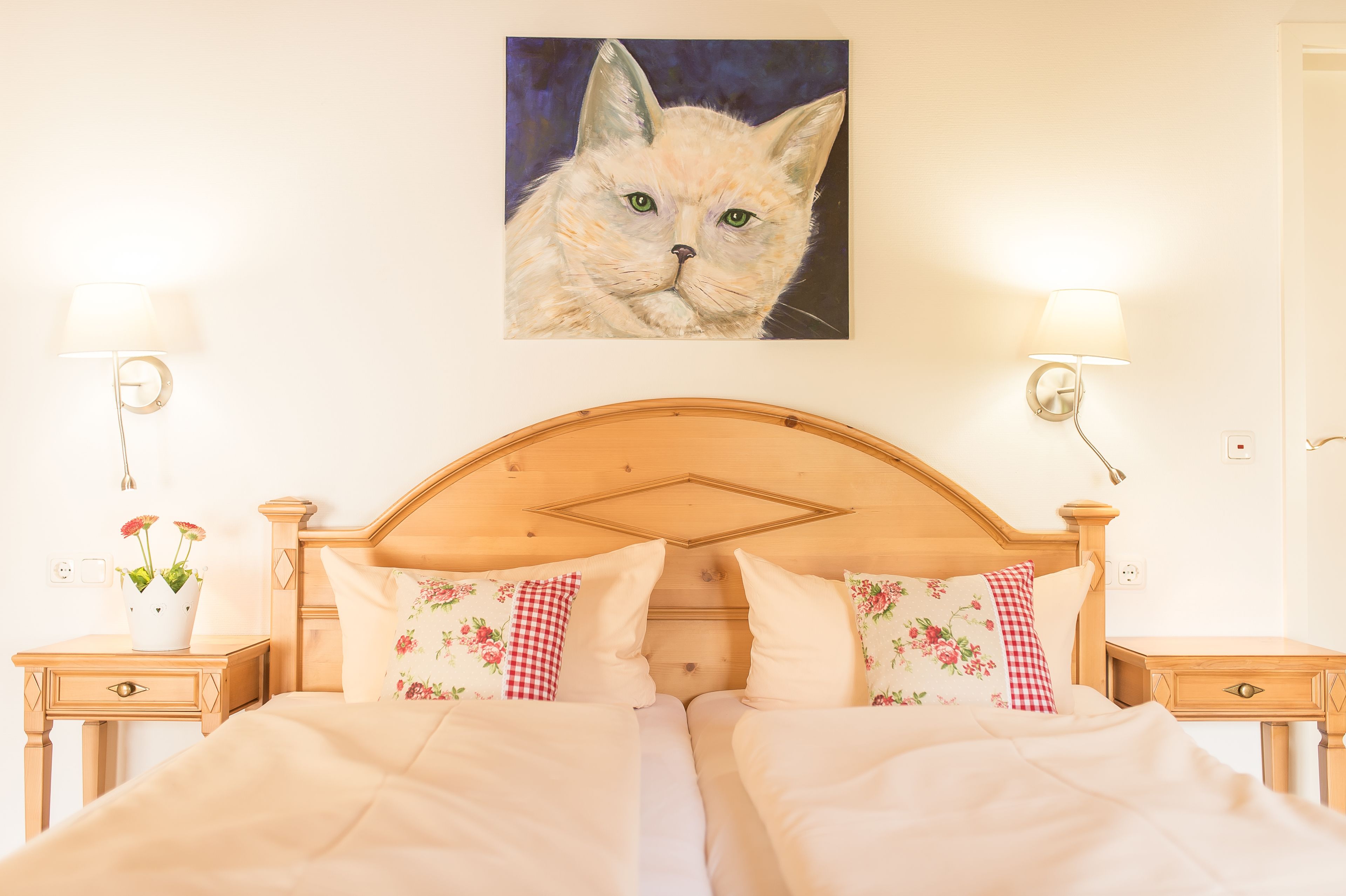 A neatly made bed with floral pillows, a wooden headboard, and a painting of a cat on the wall.