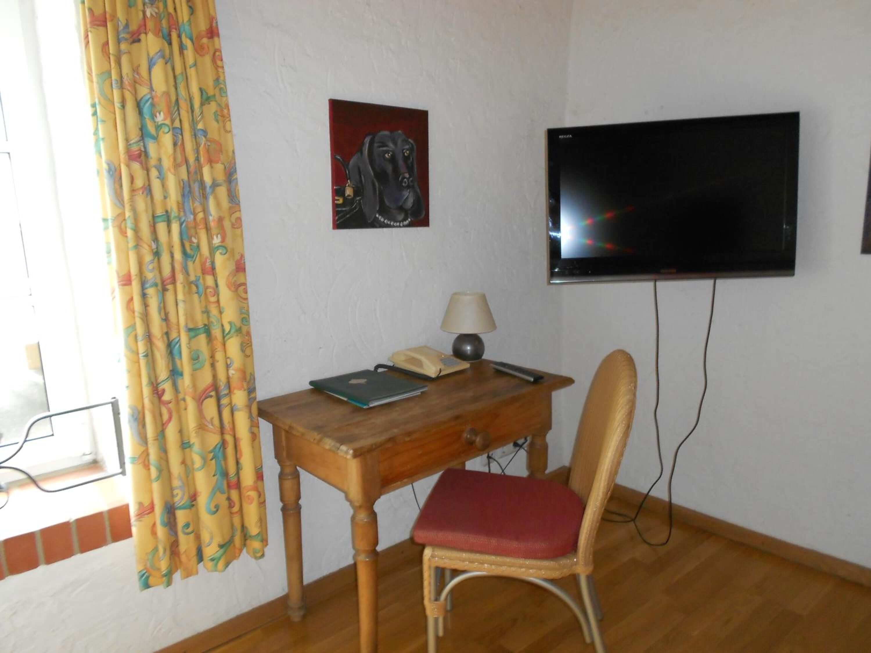 Une pièce avec un bureau en bois, une chaise, un téléviseur fixé au mur et une fenêtre avec des rideaux.