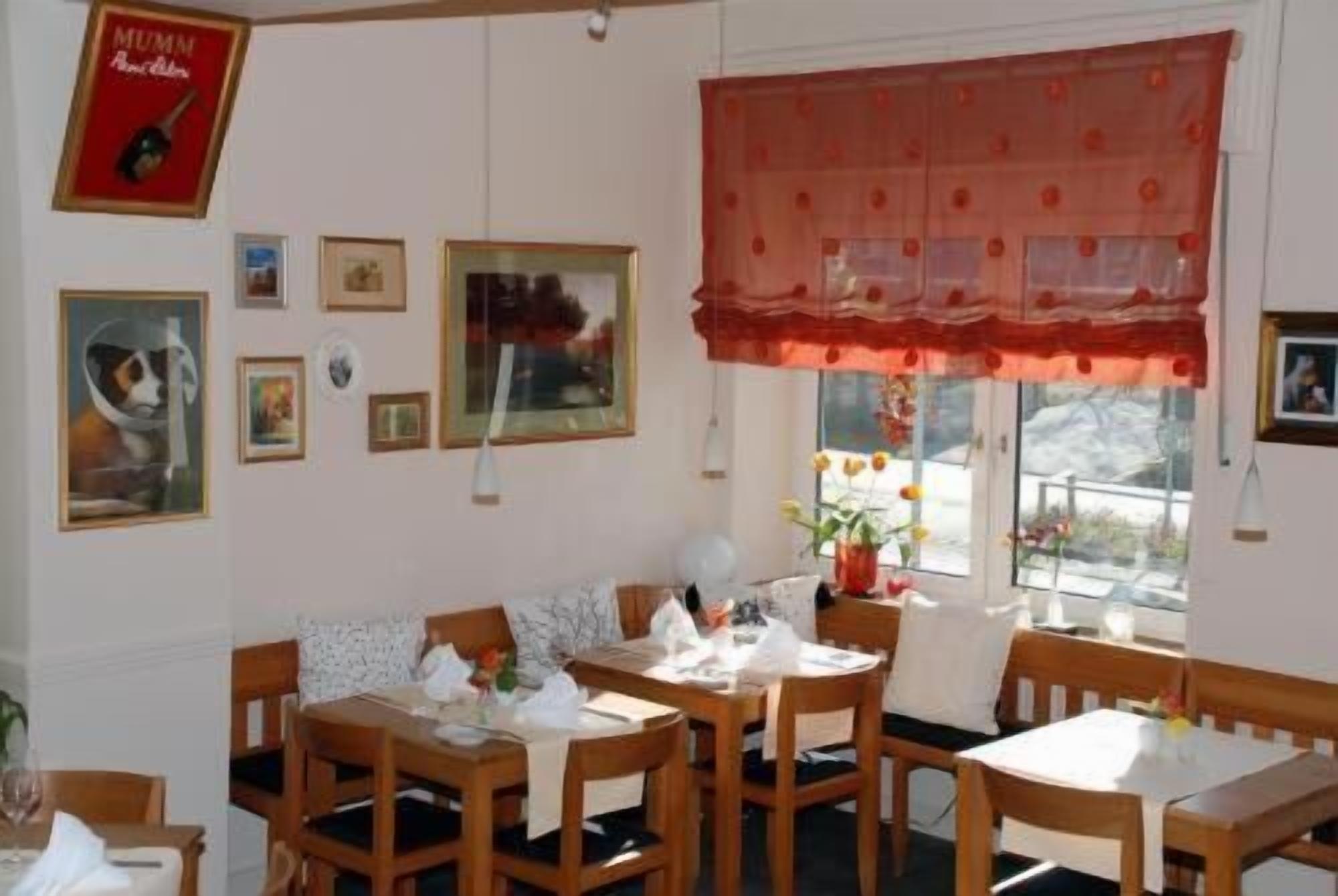 A dining area with wooden tables and chairs, framed pictures on the walls, and a window with a red valance.