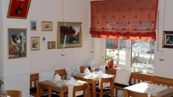 A dining area with wooden tables and chairs, framed pictures on the walls, and a window with a red valance.