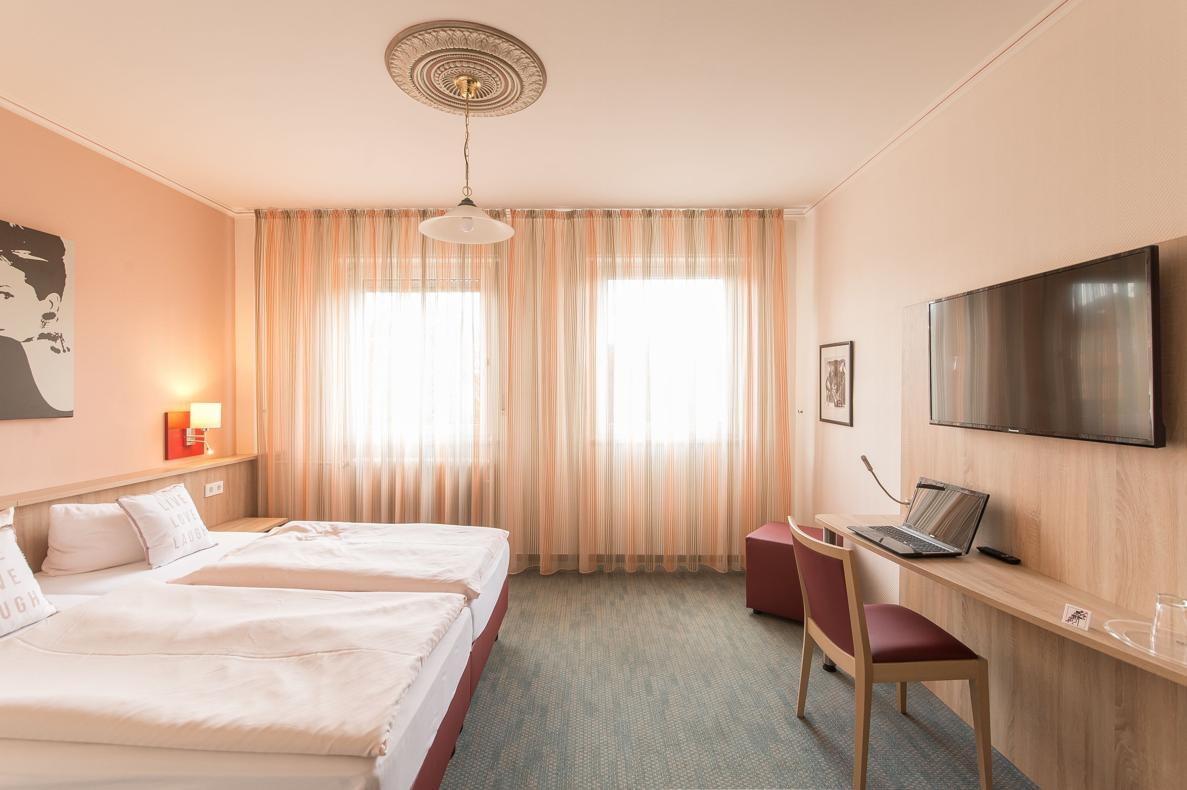 Una camera d'albergo con due letti, una scrivania con un portatile, una TV e un'ampia finestra con tende.