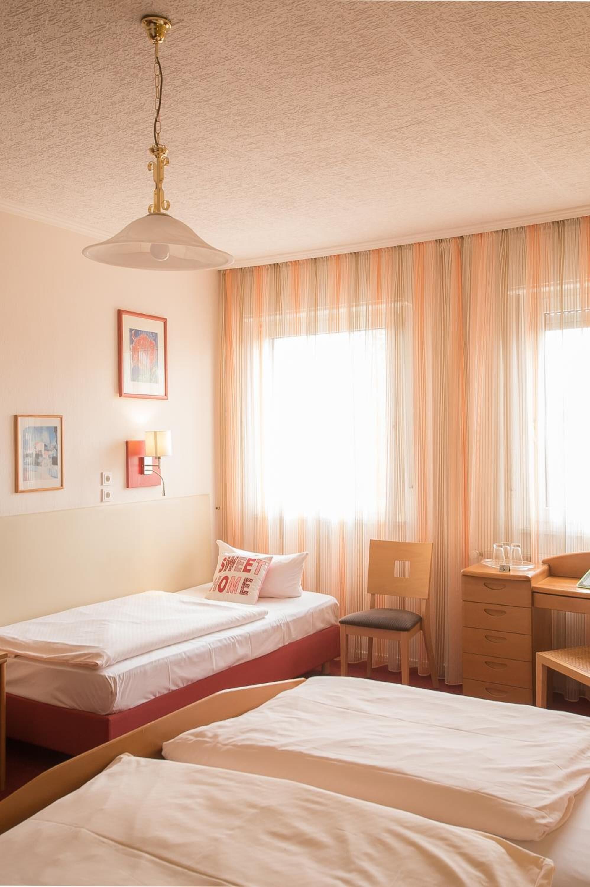 A hotel room with two single beds, a desk with a chair, a lamp, and a ceiling fan.