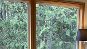 Una habitación acogedora con un ventanal grande que da a un bosque frondoso. Hay una mesita de madera con un florero y una taza. Un plaid estampado y una revista están sobre un sofá.