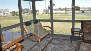 Uma varanda coberta com uma cadeira de balanço, uma mesa pequena e vista para uma área gramada com um corpo d'água e edifícios ao fundo.