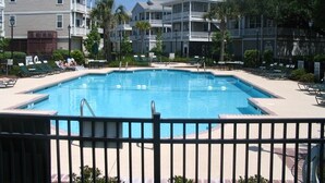 Piscina cercada por espreguiçadeiras e uma grade, com edifícios altos ao fundo.