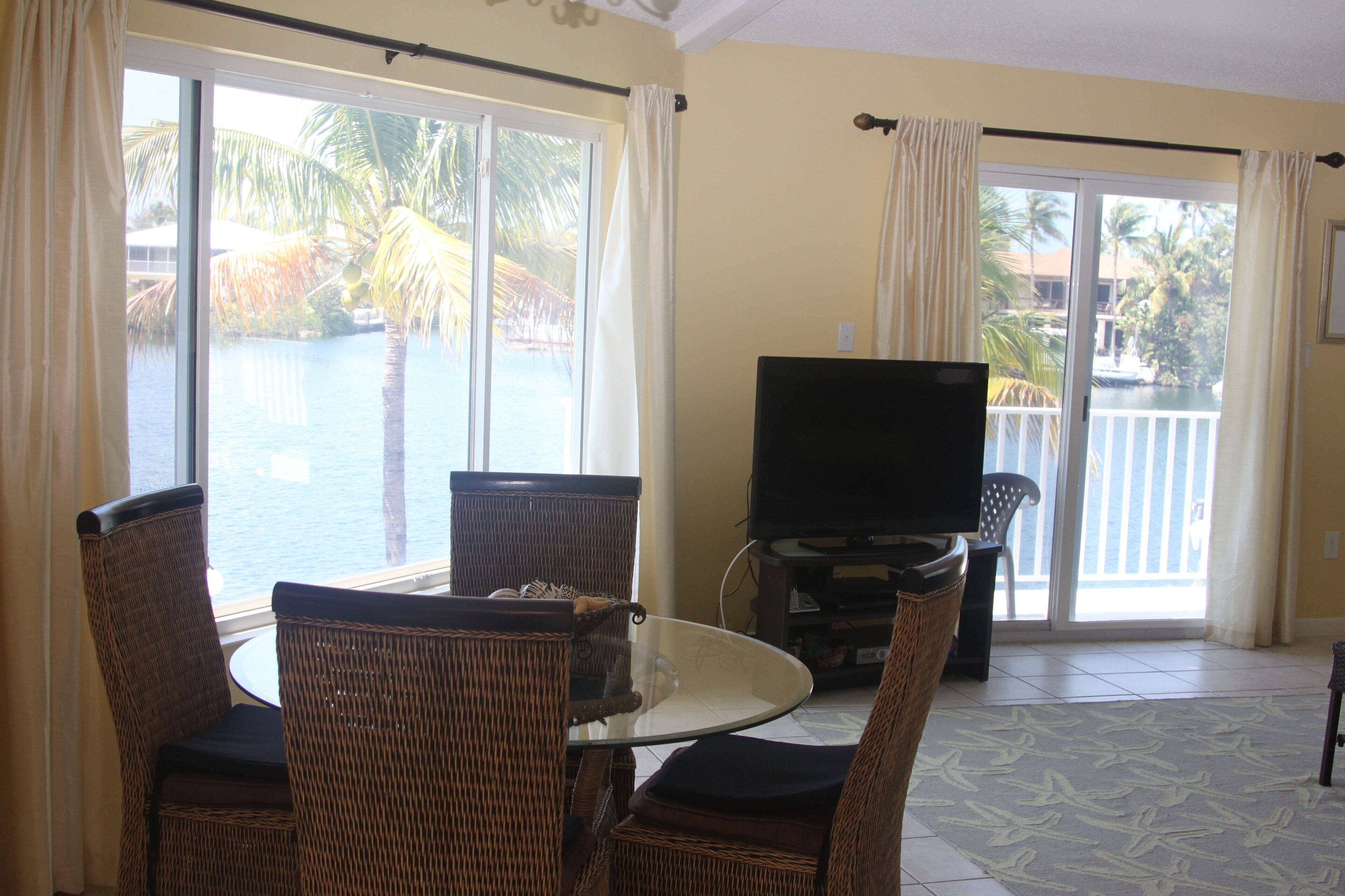 Una habitación con mobiliario de mimbre, una mesa de vidrio, un televisor de pantalla plana y vistas a una piscina y palmeras a través de amplios ventanales.
