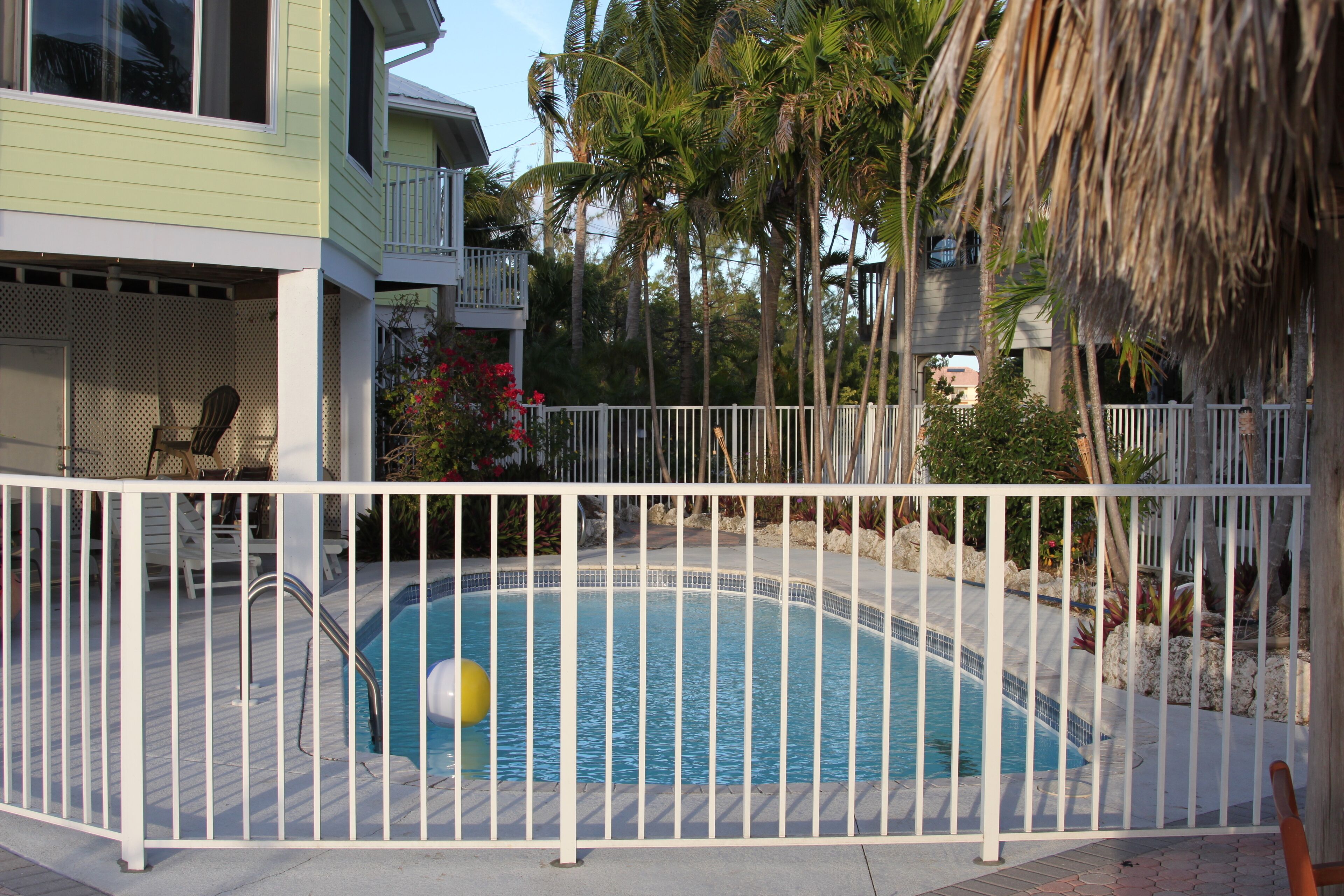 A swimming pool surrounded by a white fence, with a chair and a lounge chair nearby.