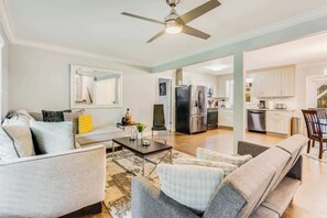 Una sala de estar moderna con un sofá, sillones y una mesa de centro. Hay un ventilador de techo, una cocina al fondo y un comedor con sillas.