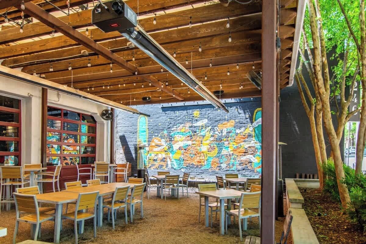 Un espace de restauration intérieur avec des tables et des chaises en bois, un mur de briques orné de graffitis colorés et un plafond avec des poutres apparentes et des luminaires suspendus.