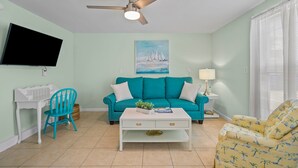 A living room with a teal sofa, a white coffee table, a blue chair, a TV mounted on the wall, and a ceiling fan. - "Sand Dollar Suites" suite 4: A short stroll from our sandy shores! (Carolina Beach)