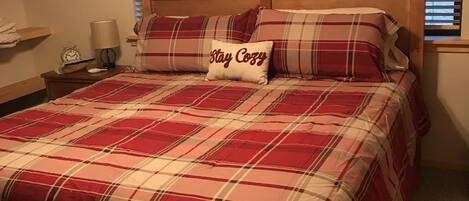 Ein Schlafzimmer mit karierter Bettdecke, einem Kissen mit dem Aufdruck 'Stay Cozy', einem Holz-Kopfteil, einem Nachttisch mit Lampe und einem Fenster mit Jalousien.