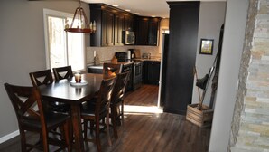 A modern kitchen with dark cabinetry, a central island, and a dining area with wooden chairs. - 4 Bedroom, 2 Bath  Wooded Getaway (Boyne Falls)