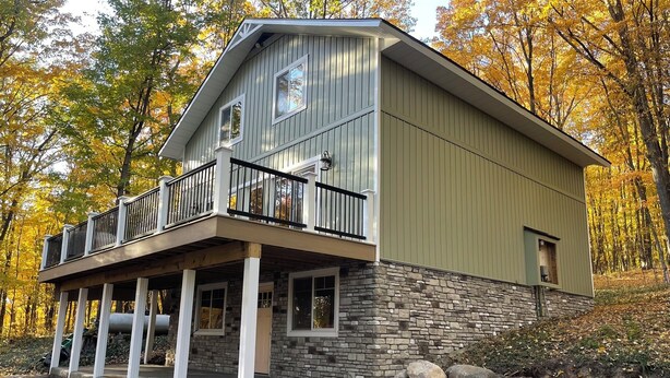 A two-story cabin with a stone foundation and a green exterior, featuring a balcony with black railings. - 4 Bedroom, 2 Bath  Wooded Getaway (Boyne Falls)