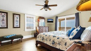 A bedroom with a large bed, a wooden bench, a ceiling fan, a TV, and a window with a view.