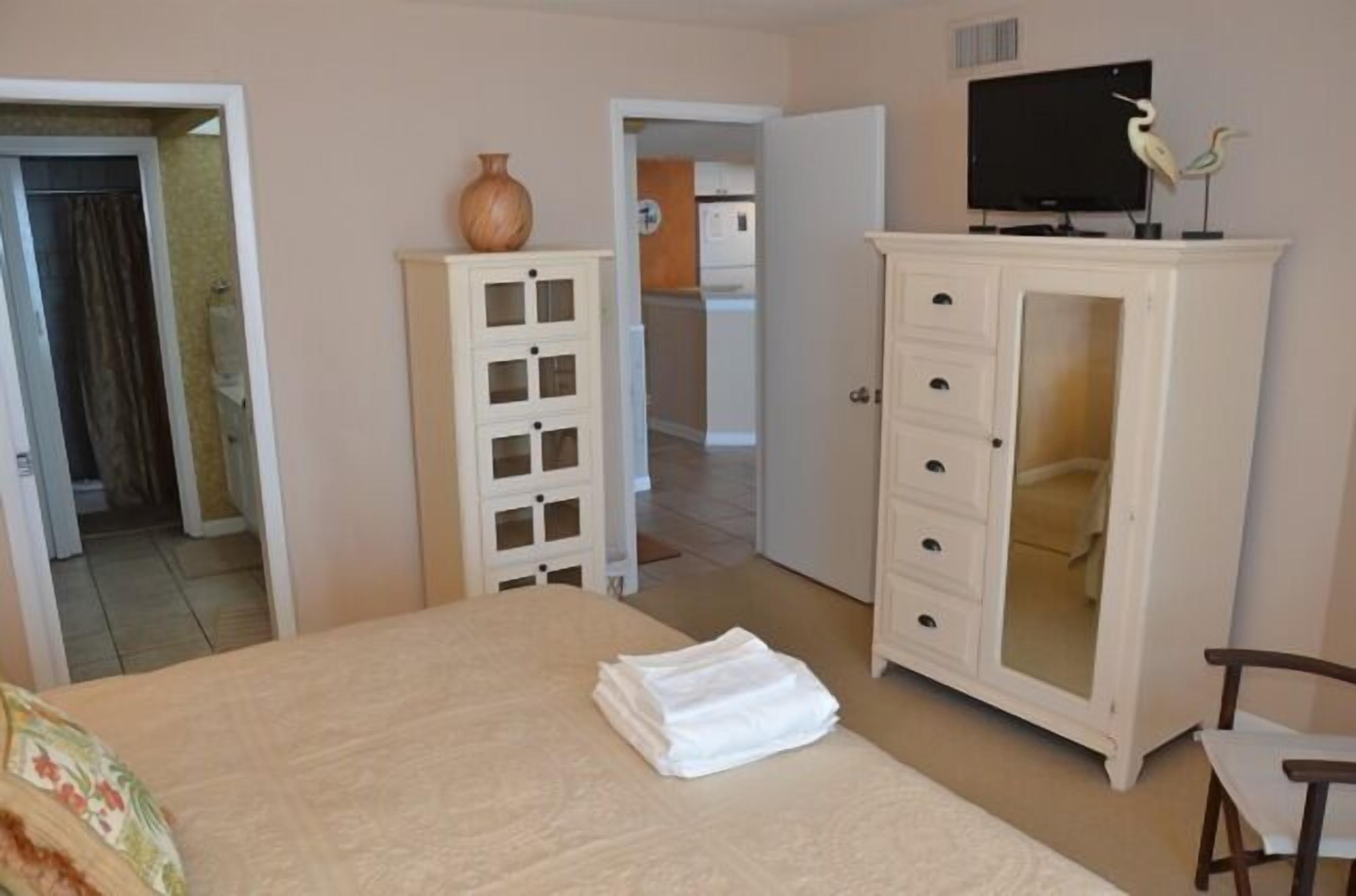 Quarto de hotel com uma cama, um armário branco com gavetas, uma televisão em um suporte e um vaso sobre o armário.