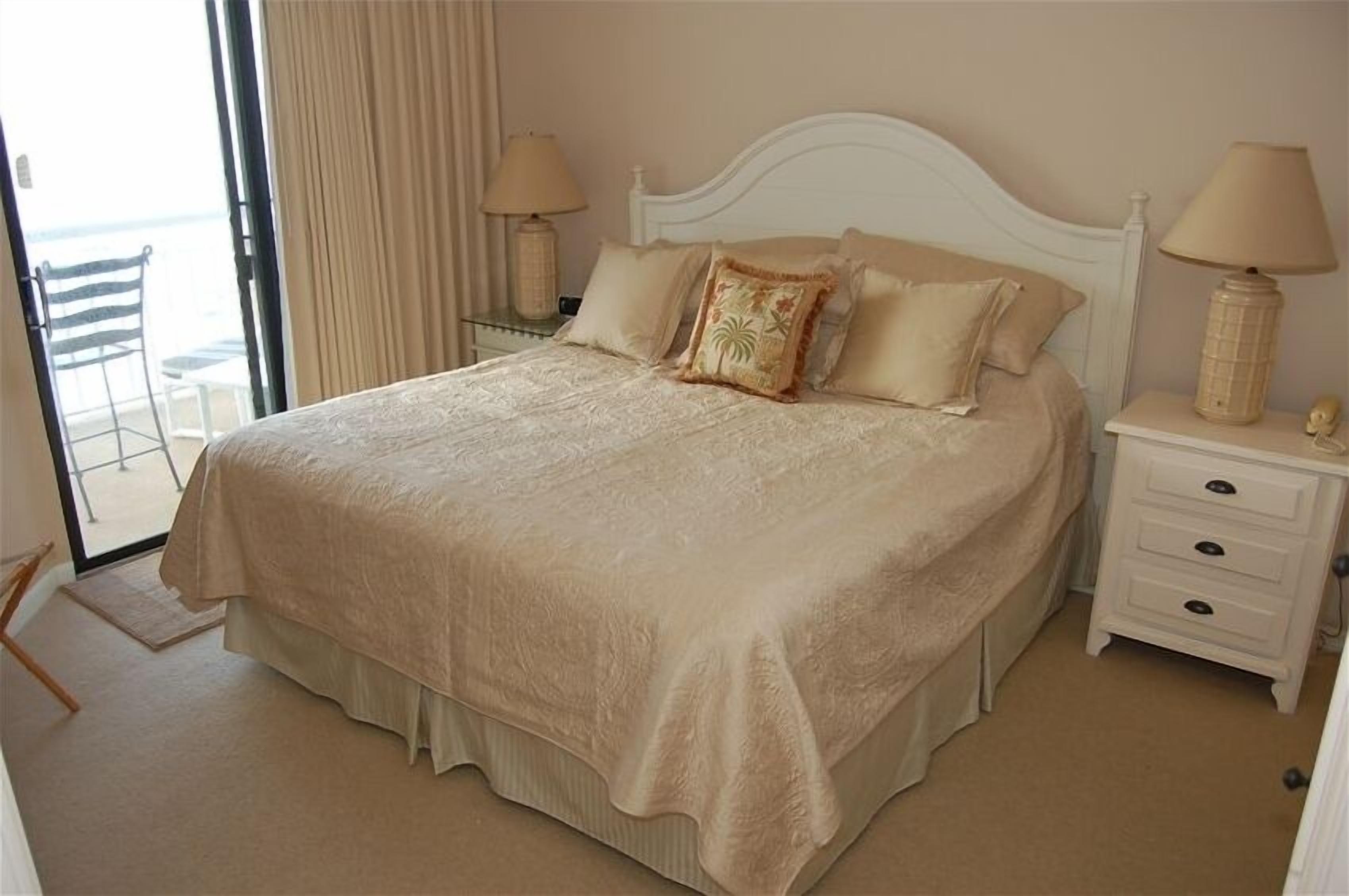A large bed with a beige bedspread and pillows, a white nightstand with a lamp, and a view of the beach through an open door.