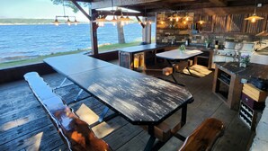 An outdoor kitchen with a view of the water, wooden furniture, and hanging lights. - Kemp's Rentals Luxurious Executive Retreat Portage Lake-Beach-Firepit & Pavilion (Hancock)