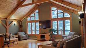 A spacious living room with a high ceiling, wooden beams, large arched windows, a flat-screen TV, and a cozy seating area. - Kemp's Rentals Luxurious Executive Retreat Portage Lake-Beach-Firepit & Pavilion (Hancock)