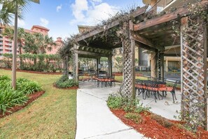 Un espace extérieur aménagé en terrasse, avec des tables et des chaises, entouré d’un jardin où poussent différentes plantes et surmonté d’une structure en pergola en bois.