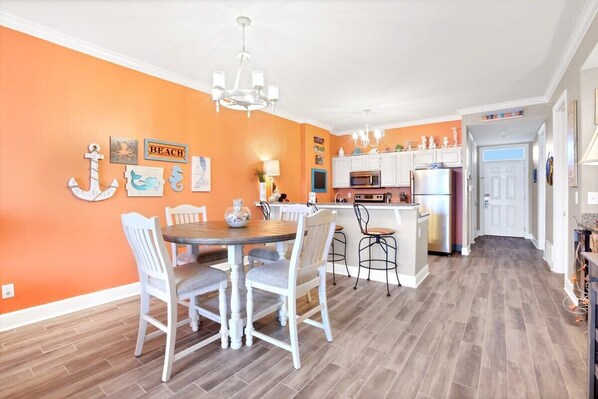 Une pièce aux murs d’un orange vif, comprenant une table à manger et des chaises, un coin cuisine avec des meubles blancs et un lustre.