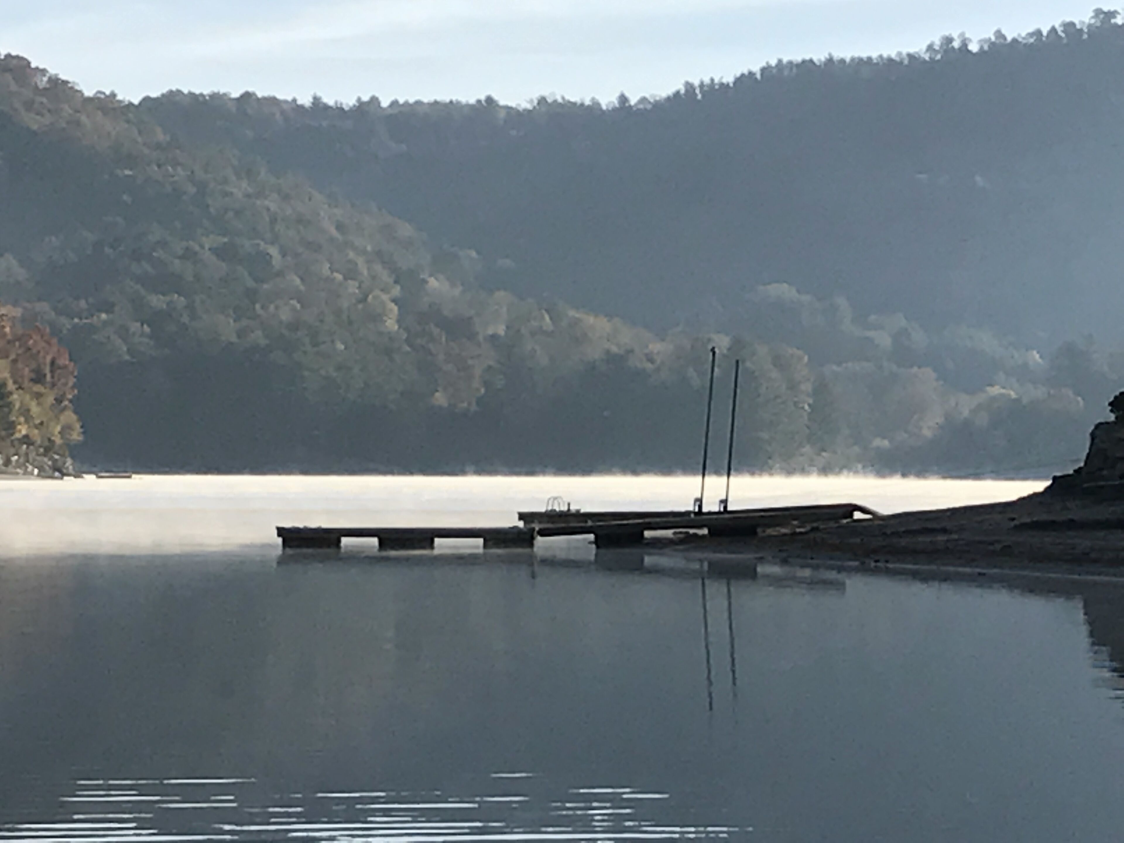 A calm lake with a dock extending into the water, surrounded by forested hills.