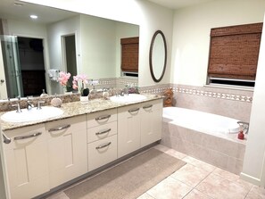 Une salle de bain avec un meuble-lavabo à deux vasques, un grand miroir, une baignoire et une fenêtre avec des stores.
