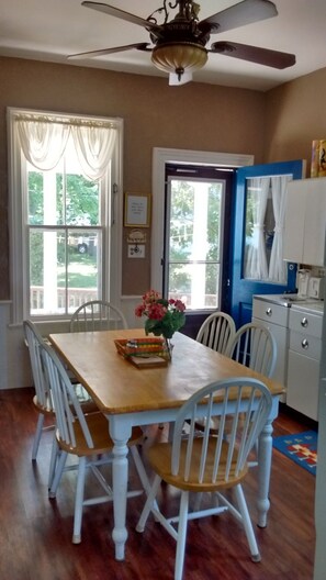 Un espace repas avec une table en bois et des chaises blanches, un ventilateur de plafond et une vue sur l’extérieur grâce à de grandes fenêtres.