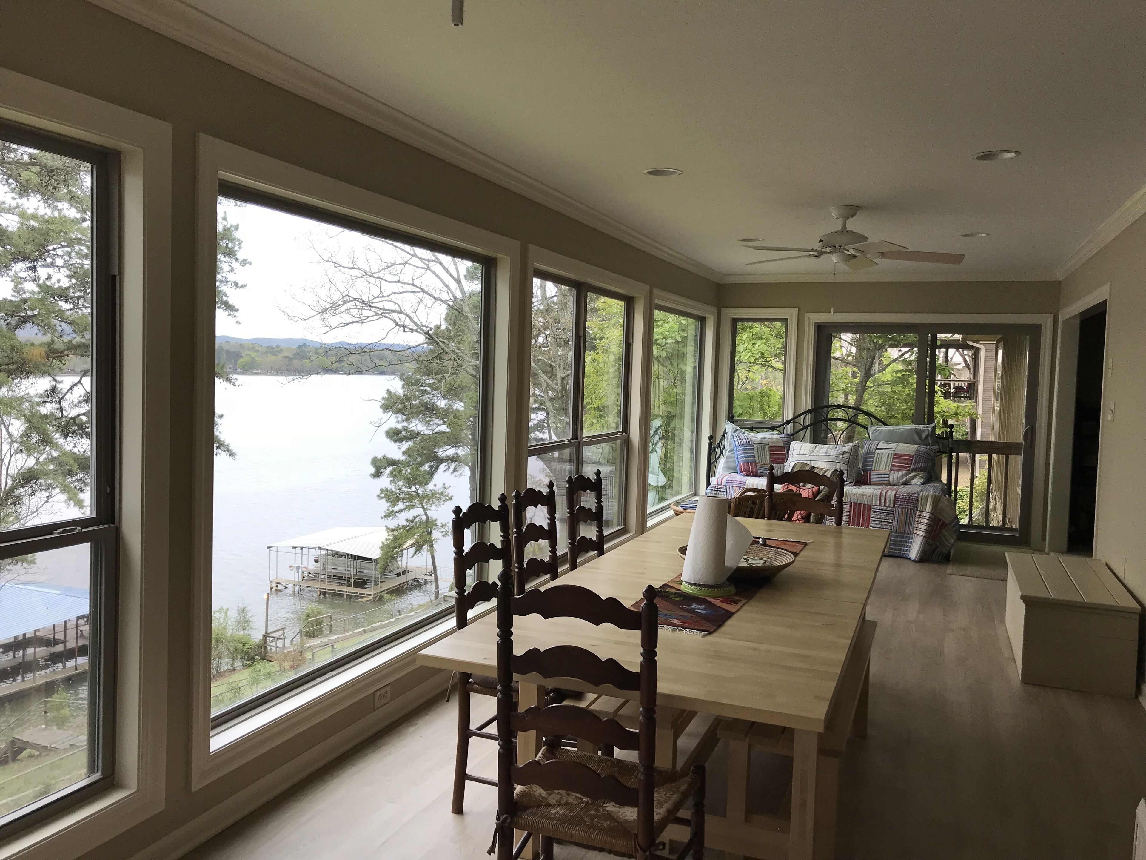A spacious room with large windows overlooking a lake, a dining area with a wooden table and chairs, and a cozy seating area with a bed and pillows.