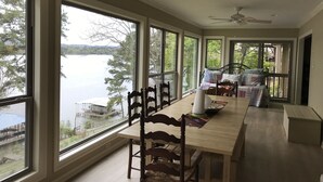 Een ruime kamer met grote ramen met uitzicht op een meer, een eethoek met een houten tafel en stoelen, en een gezellige zithoek met een bed en kussens.