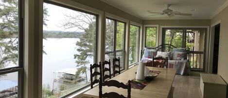 A spacious room with large windows overlooking a lake, a dining area with a wooden table and chairs, and a cozy seating area with a bed and pillows.
