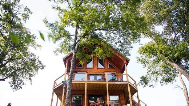 A two-story wooden cabin with a balcony, surrounded by trees and grass.