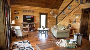A cabin interior with a wooden floor, a sofa, a chair, a television, and a staircase.