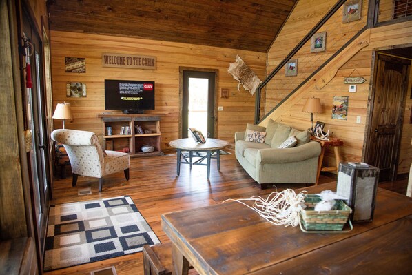 A cabin interior with a wooden floor, a sofa, a chair, a television, and a staircase.