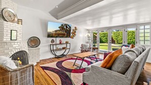 Amplia sala de estar con chimenea, un televisor de pantalla plana grande, un sofá cómodo y vistas a un jardín exuberante a través de amplios ventanales.