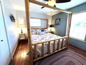 A bedroom with a wooden four-poster bed, a window with blinds, a nightstand with a lamp, and a wall-mounted picture.