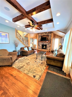 A spacious living room with a stone fireplace, wooden beams, and a ceiling fan.