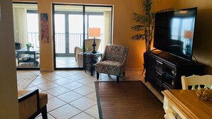 A living room with a TV, a patterned armchair, a dining table, and a large window. - Seabatical at The Gulf Tower, Beautiful Beachfront Condo (Gulf Shores)