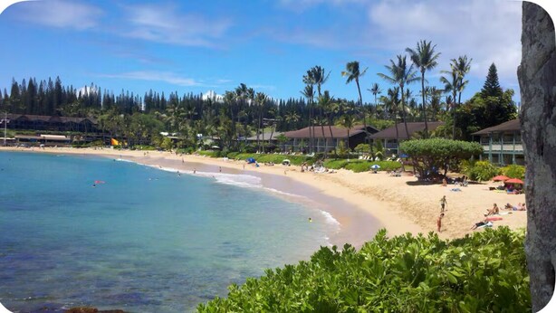A beach with clear turquoise water, sandy shore, and palm trees. There are buildings with a tropical architectural style in the background. - Beautiful Three Bedroom Townhouse at Napili Bay (Lahaina)