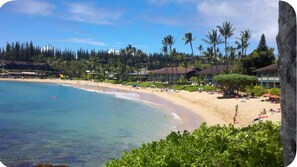 A beach with clear turquoise water, sandy shore, and palm trees. There are buildings with a tropical architectural style in the background. - Beautiful Three Bedroom Townhouse at Napili Bay (Lahaina)