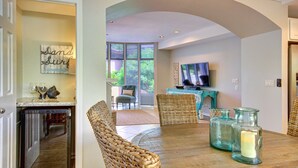 A dining area with a wooden table, wicker chairs, a television, and a kitchen with a dishwasher. - Beautiful Three Bedroom Townhouse at Napili Bay (Lahaina)