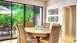 A dining area with a round table and wicker chairs, a glass door leading to a garden, and framed pictures on the wall. - Beautiful Three Bedroom Townhouse at Napili Bay (Lahaina)